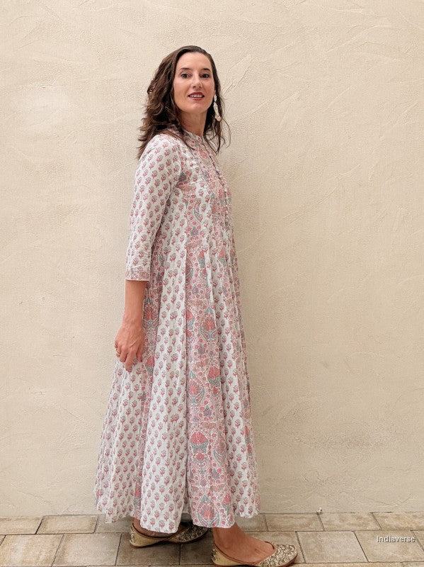 A woman modeling a long-sleeved, knee-length white kurta with pink and green floral block prints, standing against a textured beige stucco wall.