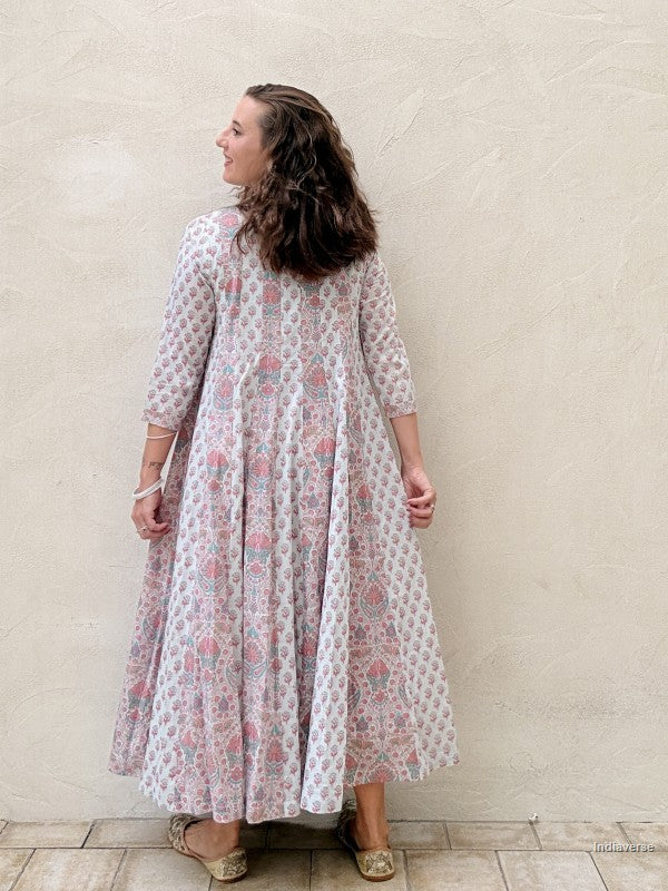 A woman wearing a long-sleeved, knee-length maxi dress with a pink and green floral block print on a white base, standing against a textured beige stucco wall.
