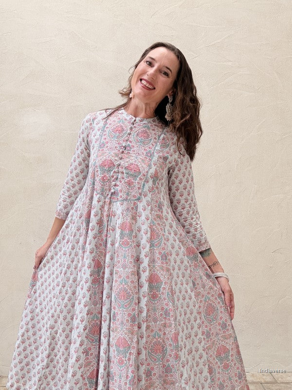 A woman wearing a long-sleeved, high-neck maxi dress with a pink and teal floral block print on a white base, standing against a textured beige wall.
