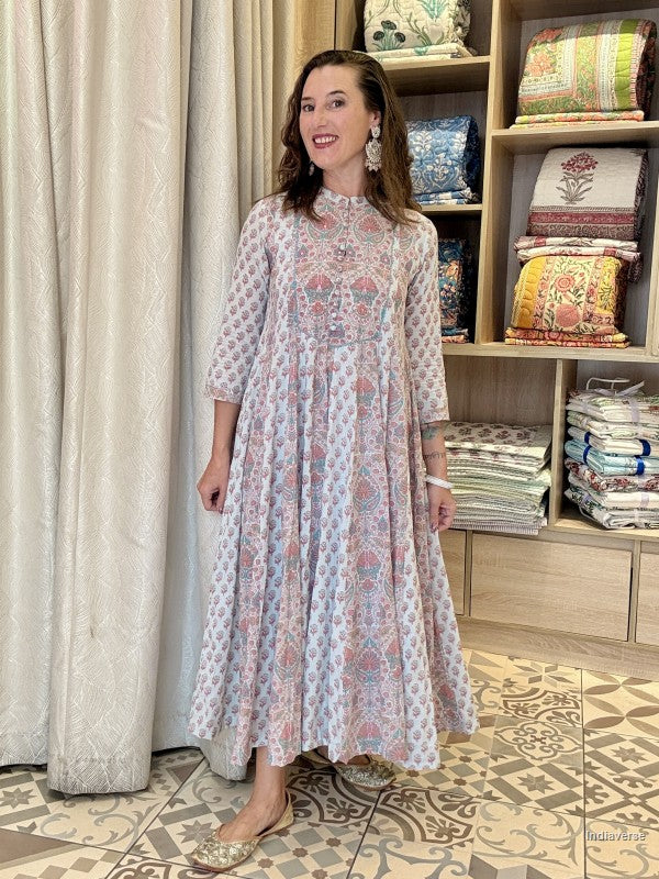 A woman modeling a long-sleeved, high-neck maxi dress with a pink, blue, and white floral block print. She is standing in a boutique setting with a patterned tile floor, a beige curtain, and wooden shelves filled with folded textiles in the background.