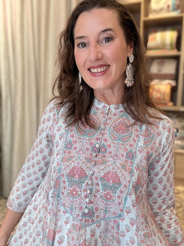 A smiling woman with brown hair is wearing a white three-quarter sleeve top with a pink and teal floral block print pattern. She is wearing large silver-toned statement earrings, and the background shows a softly blurred indoor setting with curtains and bookshelves.
