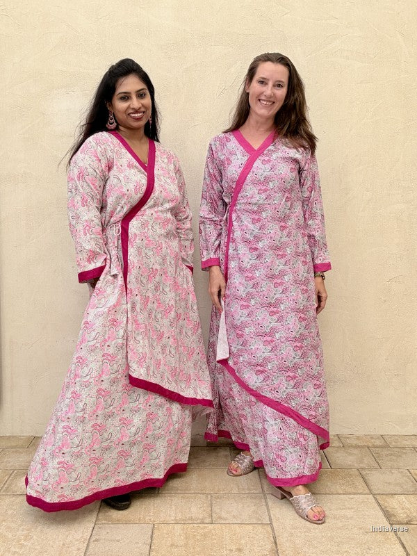 Two women wearing matching pink and white floral outfits with pink borders against a beige wall.