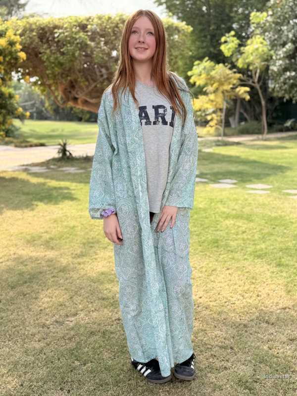 Person wearing a long light green dress over a gray sweatshirt with 'ARI' text, standing in a park.