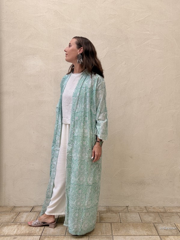 Woman wearing a long light green floral dress against a beige wall.