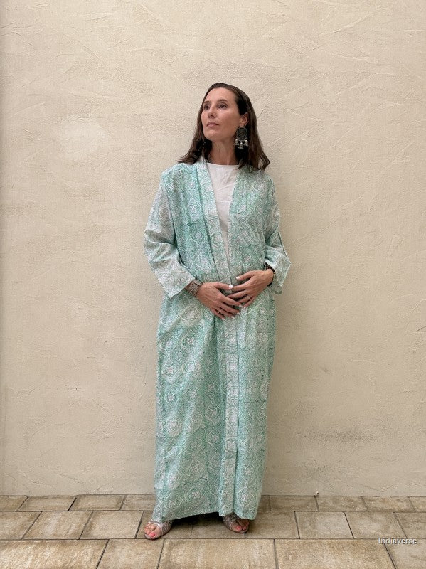 Woman wearing a long, light green dress with a pattern against a beige wall.