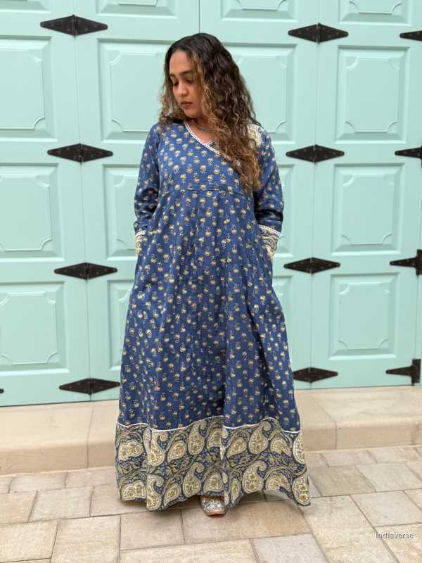 woman wearing royal blue colored festive wrap dress with silver border neckline and paisley borders at the trim and sleeves in front of a light blue door