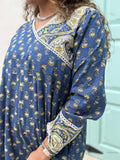 close up of woman wearing royal blue colored festive wrap dress with silver border neckline and paisley borders at the trim and sleeves in front of a light blue door