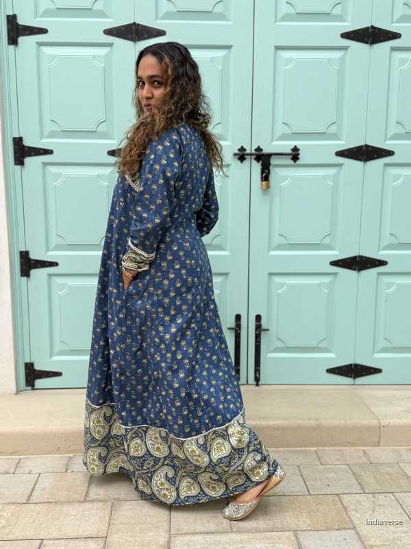 woman wearing royal blue colored festive wrap dress with silver border neckline and paisley borders at the trim and sleeves in front of a light blue door