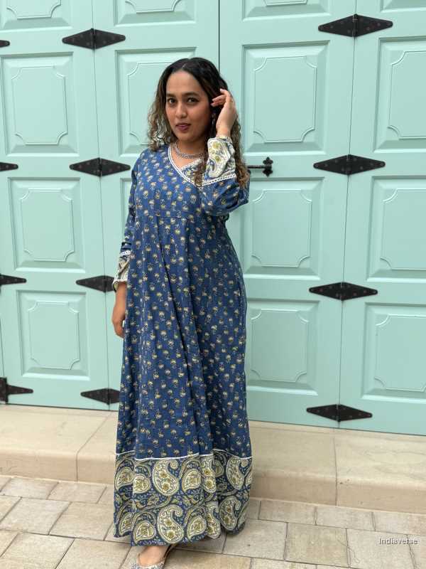 woman wearing royal blue colored festive wrap dress with silver border neckline and paisley borders at the trim and sleeves in front of a light blue door