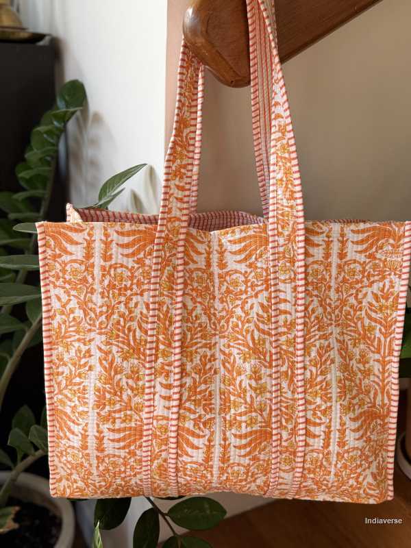 Orange and white patterned tote bag hanging on a wooden surface with plants in the background.
