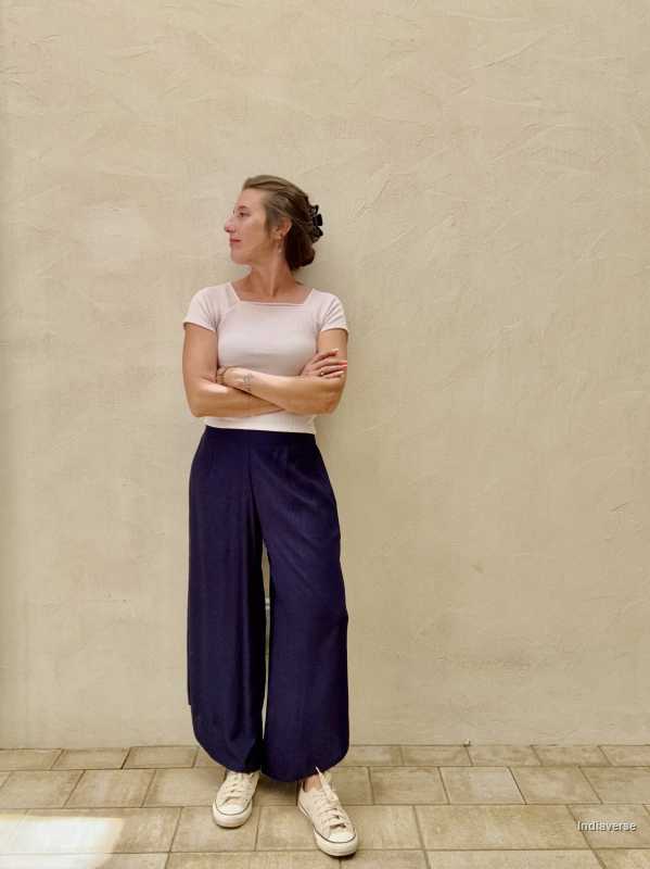 A woman wearing a short top and blue palazzo pants is standing against a beige wall