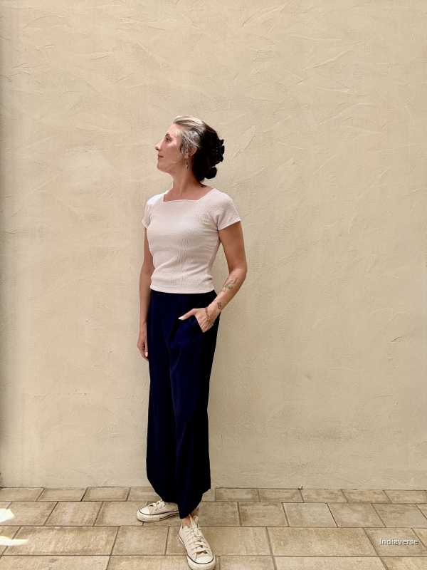 A woman wearing a light color top and dark blue palazzo pants is standing against a beige wall