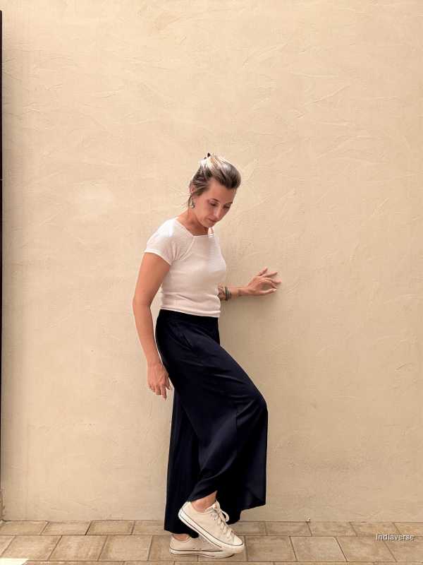 Woman in a white top and dark blue pants leaning against a beige wall.