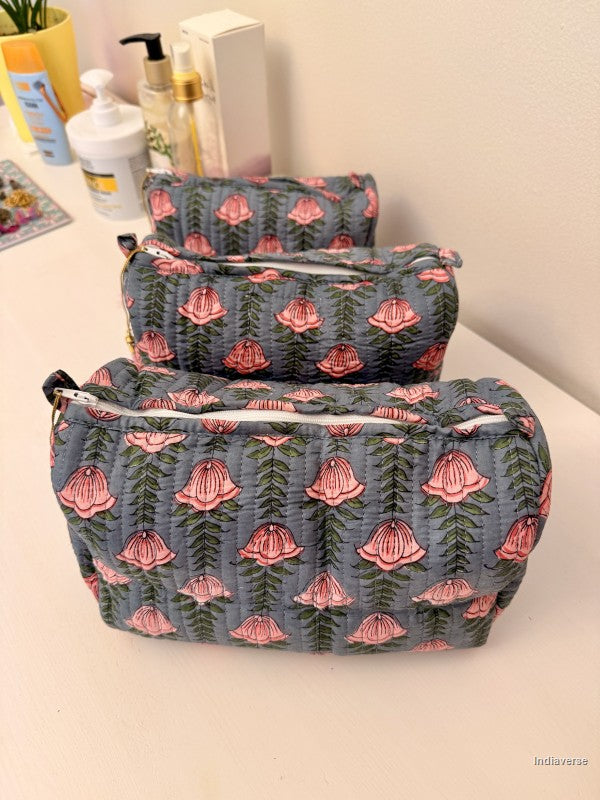 Three quilted fabric cosmetic pouches in a tiered stack, featuring a blue-gray background with a pink and green floral bell pattern, set on a light-colored tabletop | Pink Lotus