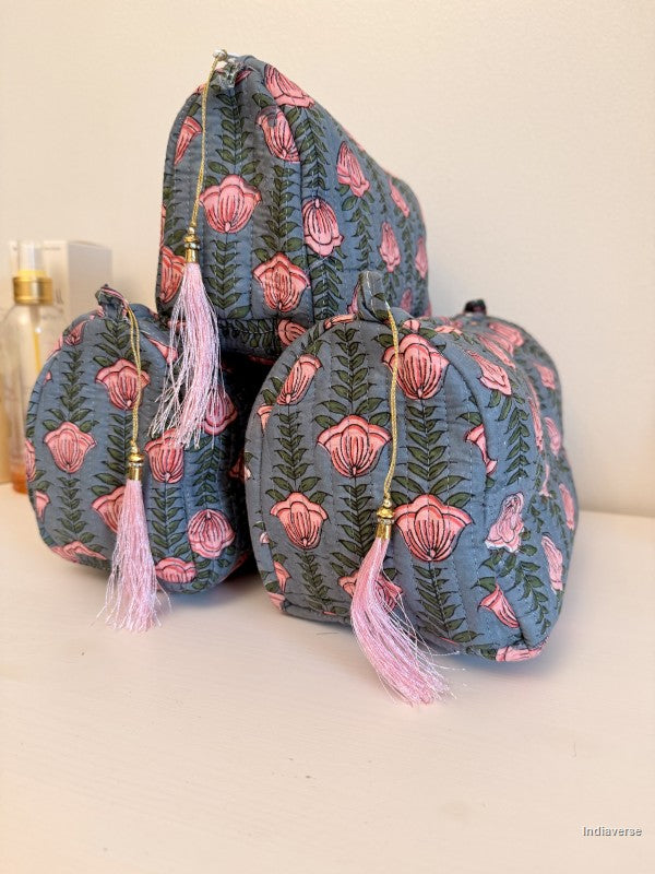 Three small zippered cosmetic pouches in a dusty blue fabric with a pink and green floral print, decorated with light pink tassels and gold zipper pulls, arranged on a white surface | Pink Lotus