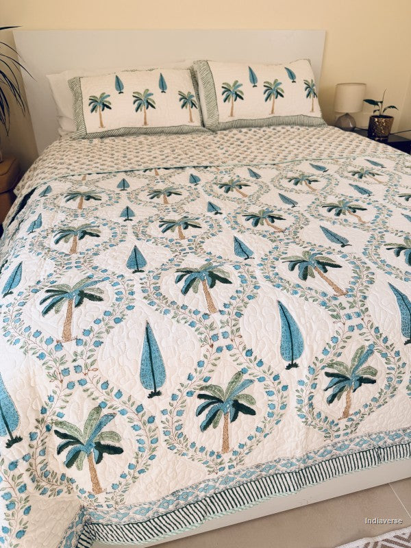 A tropical-themed quilted bedspread and matching pillow shams featuring embroidered palm trees and blue leaves on an off-white background in a well decorated bedroom.