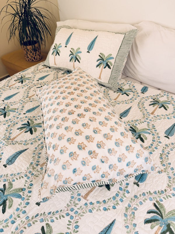 A tropical-themed quilted bedspread and matching pillow shams featuring embroidered palm trees and blue leaves on an off-white background nicely arranged on a queen size bed