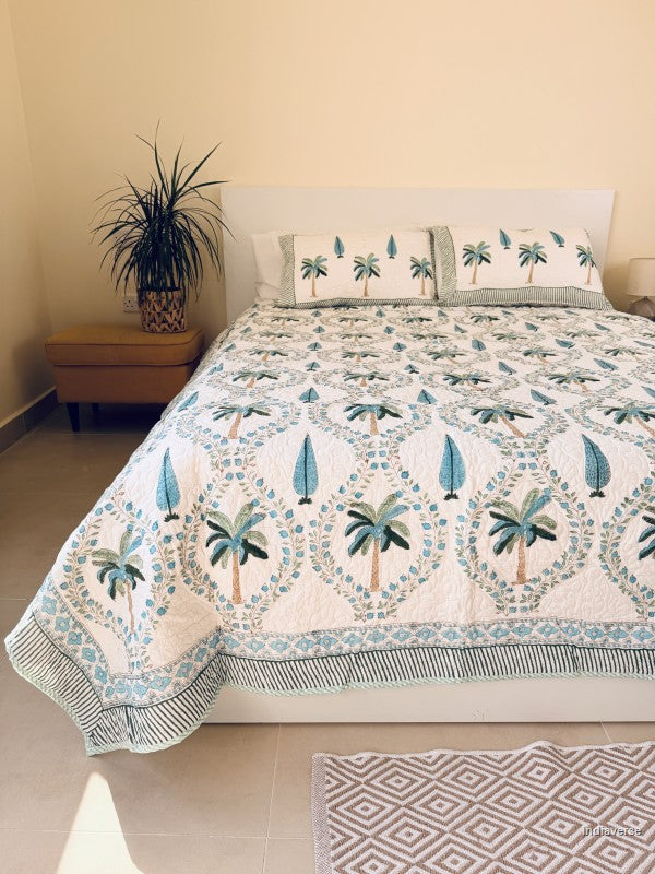 A tropical-themed quilted bedspread and matching pillow shams featuring embroidered palm trees and blue leaves on an off-white background nicely arranged on a queen size bed in a beige bedroom