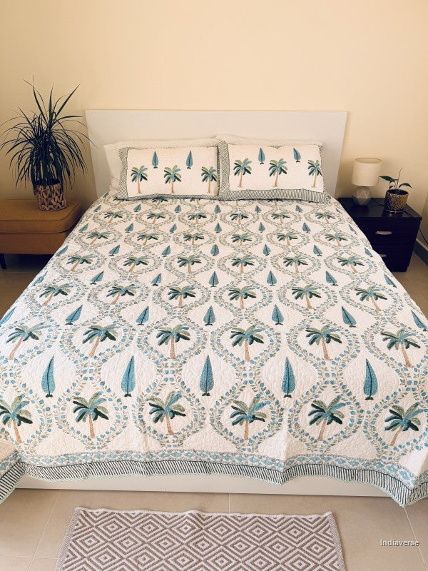 A tropical-themed quilted bedspread and matching pillow shams featuring embroidered palm trees and blue leaves on an off-white background neatly arranged on a queen size bed in a warm decorated bedroom