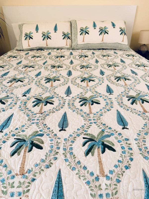 A tropical-themed quilted bedspread and matching pillow shams featuring embroidered palm trees and blue leaves on an off-white background arranged neatly on a queen size bed.
