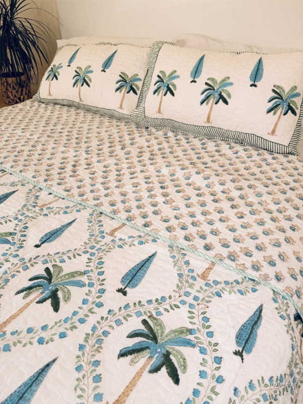 A tropical-themed quilted bedspread and matching pillow shams featuring embroidered palm trees and blue leaves on an off-white background.