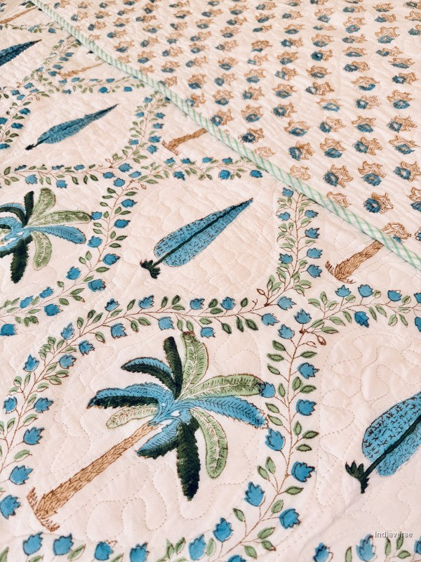 Close-up of a quilted fabric featuring embroidered palm trees, blue flowers, and leafy vines on a light cream background.