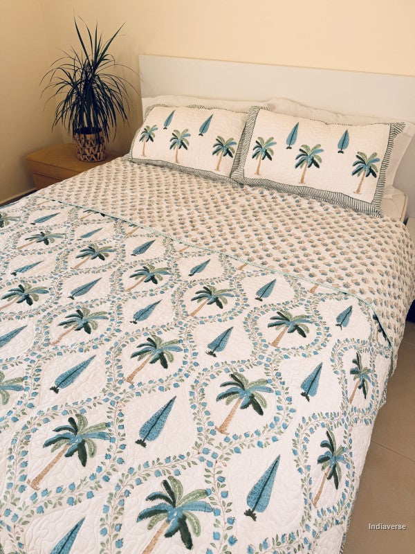 A tropical-themed quilted bedspread and matching pillow shams featuring embroidered palm trees and blue leaves on an off-white background draped on a queen size bed.