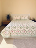 A quilted bedspread and matching pillow shams featuring a tropical pattern of green palm trees and pink flowers on an off-white background arranged neatly on a queen size bed