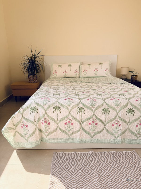 A quilted bedspread and matching pillow shams featuring a tropical pattern of green palm trees and pink flowers on an off-white background arranged neatly on a queen size bed