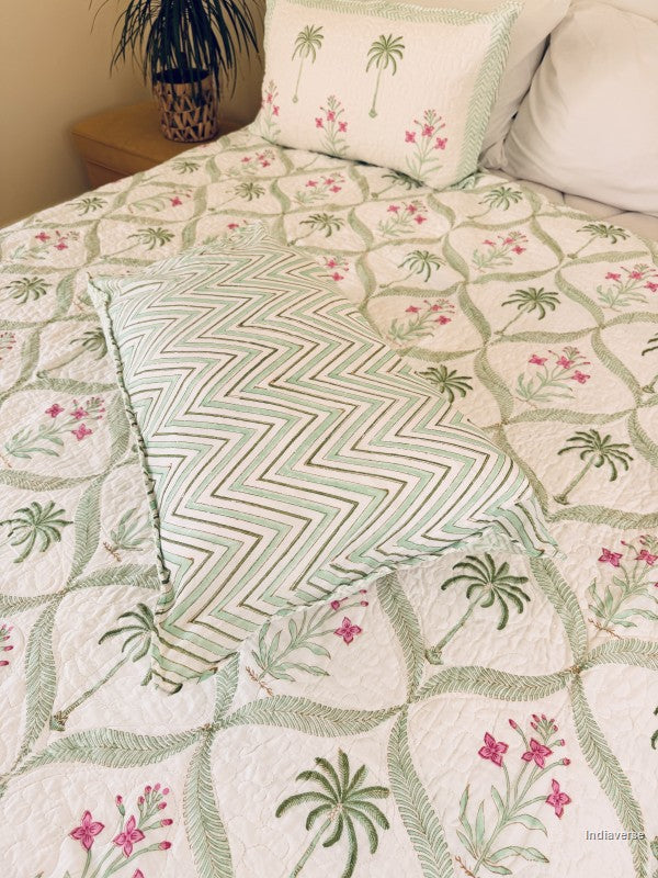 A quilted bedspread and matching pillow shams featuring a tropical pattern of green palm trees and pink flowers on an off-white background.