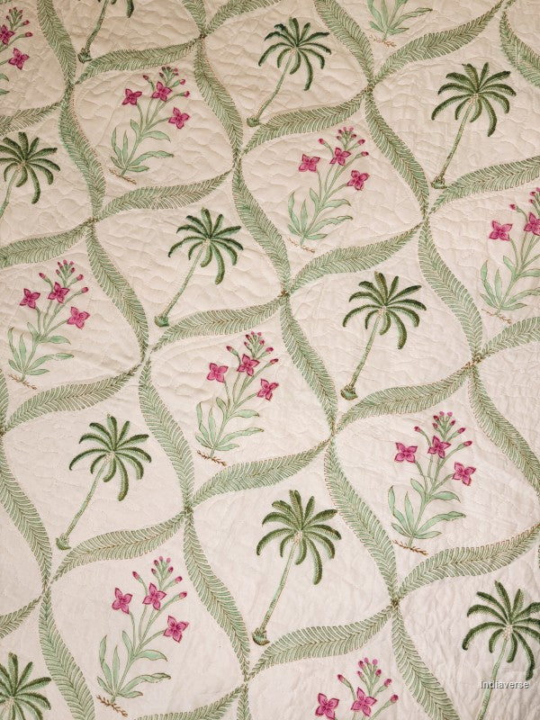 Close up of a quilted bedspread featuring a tropical pattern of green palm trees and pink flowers on an off-white background.