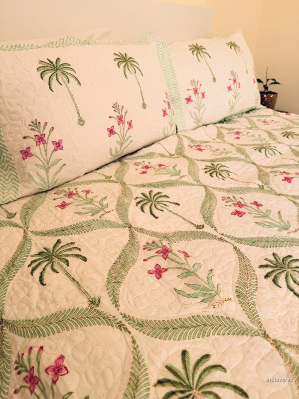 A quilted bedspread and matching pillow shams featuring a tropical pattern of green palm trees and pink flowers on an off-white background.