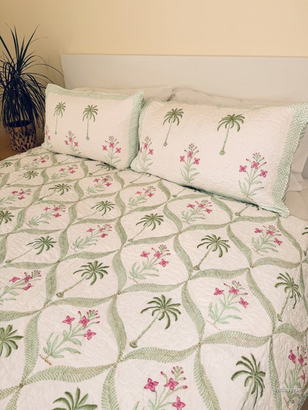A quilted bedspread and matching pillow shams featuring a tropical pattern of green palm trees and pink flowers on an off-white background placed on a queen size bed