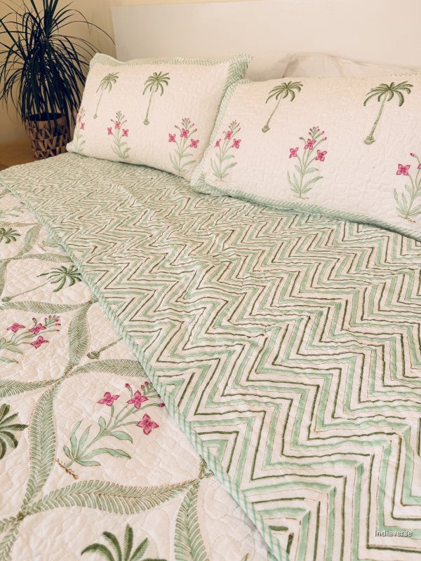 A quilted bedspread showing front and reverse side and matching pillow shams featuring a tropical pattern of green palm trees and pink flowers on an off-white background.