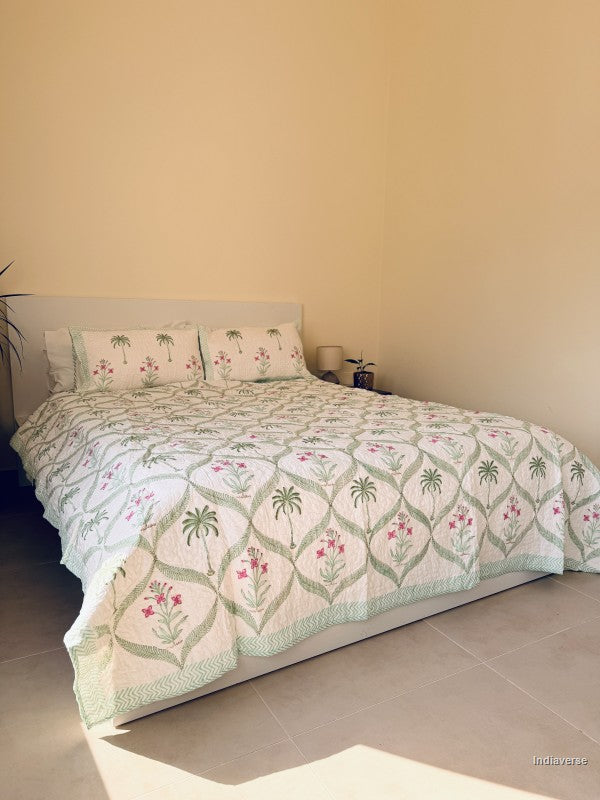 A quilted bedspread and matching pillow shams featuring a tropical pattern of green palm trees and pink flowers on an off-white background arranged on a queen size bed in a well lit bedroom