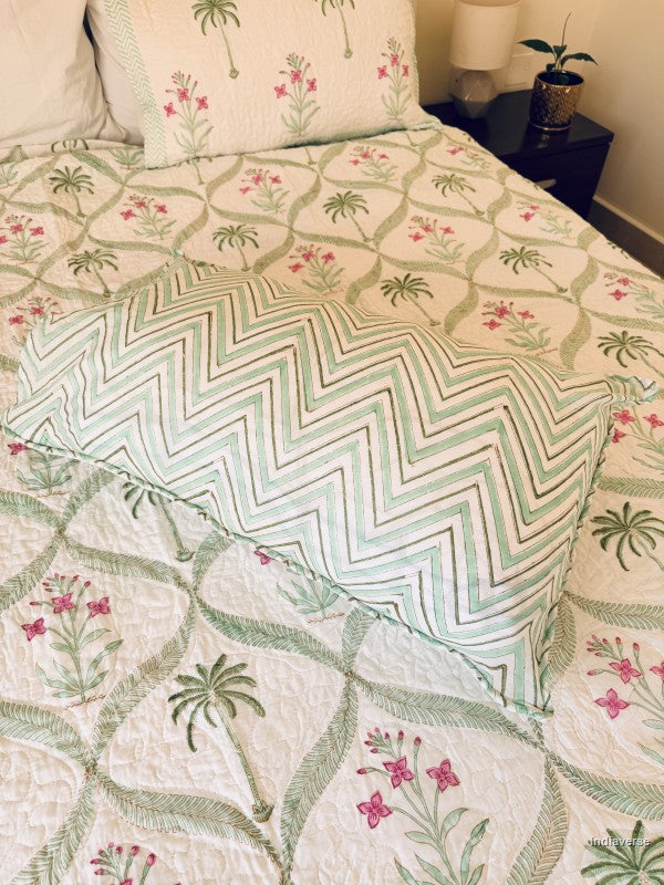 A quilted bedspread and matching pillow shams featuring a tropical pattern of green palm trees and pink flowers on an off-white background.