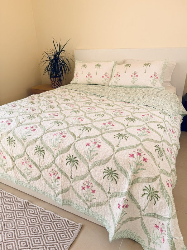 A quilted bedspread and matching pillow shams featuring a tropical pattern of green palm trees and pink flowers on an off-white background in a well decorated bedroom.