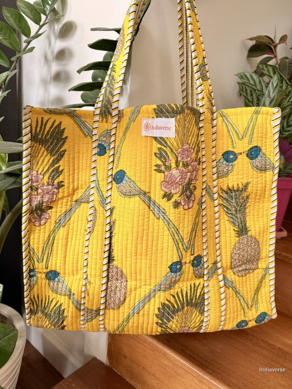 Yellow quilted tote bag with floral and bird design on a wooden surface with plants in the background.