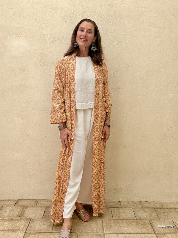 Woman wearing a long, patterned coat over a white outfit against a beige wall.