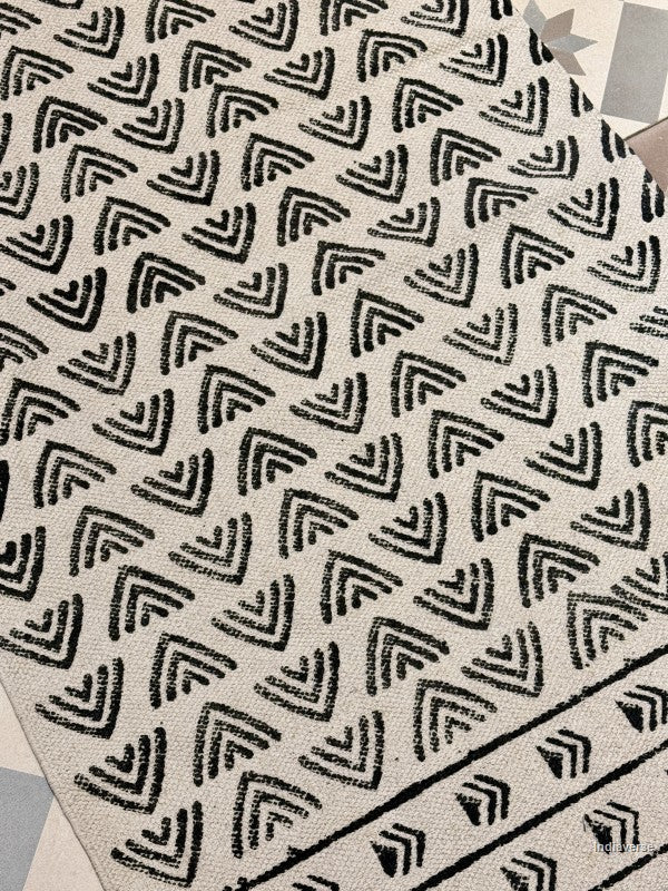 A close-up of a woven rug or textile with a repeating black geometric chevron pattern on a warm off-white background.