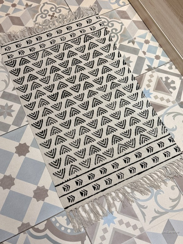 A rectangular woven runner rug with fringe, featuring a black geometric chevron and arrow pattern on a cream background, laid out on a patterned tile floor.