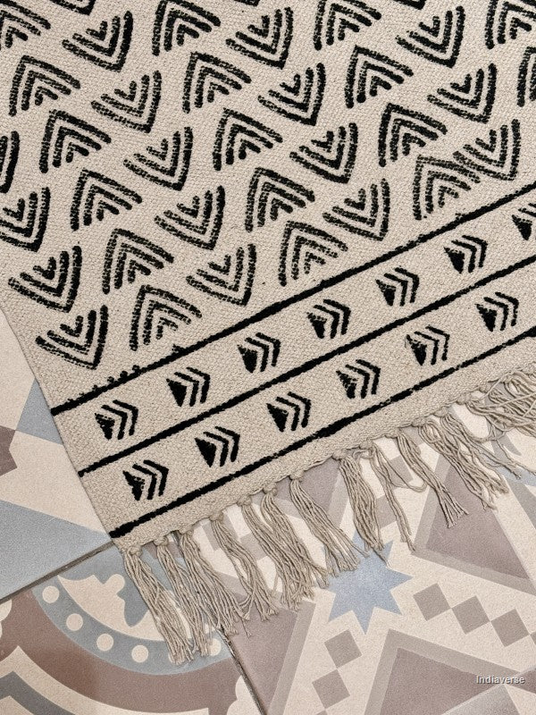 A close-up of a woven rug with a black geometric chevron and border pattern on a cream background, finished with fringe tassels, laid over a patterned tile floor.