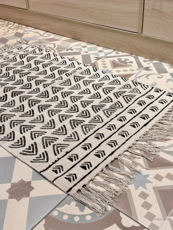 A close-up of a woven area rug with a black geometric chevron pattern on a cream background, styled on a patterned tile floor in front of a light wood cabinet.