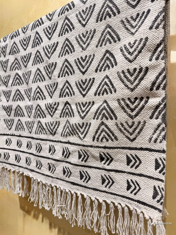 A woven textile with a black geometric chevron and arrow pattern on a light beige background, finished with white fringe along the bottom edge.