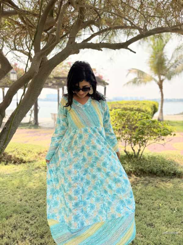 Woman in a blue and green patterned dress standing outdoors near trees and grass.
