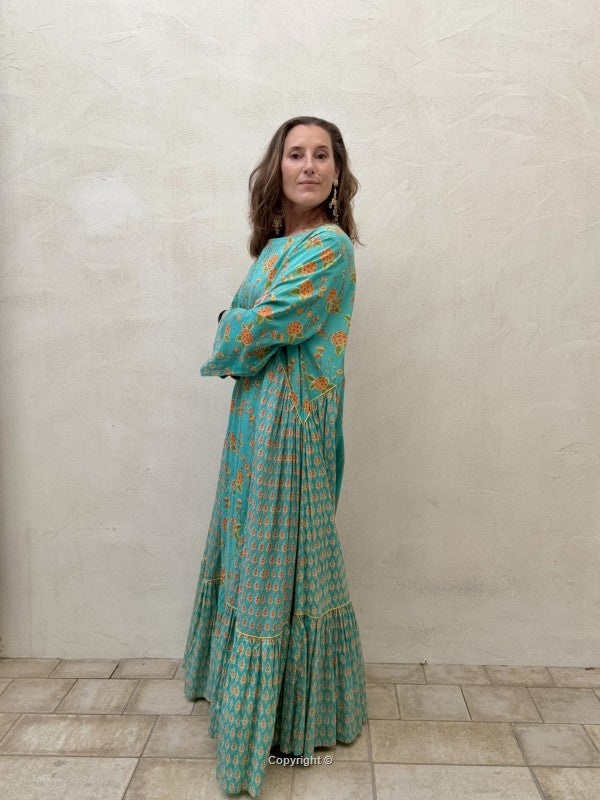 Woman wearing a teal patterned dress standing against a beige wall.